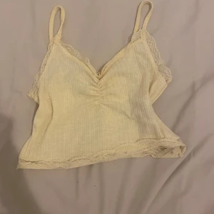 Beige ribbad linnetopp med spetsdetaljer - Söt beige linnetopp med smala axelband och spetsdetaljer längs kanterna. Toppen är ribbad och har en lätt rynkning framtill för en feminin touch. Perfekt för varma dagar eller som ett lager under en kofta.