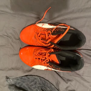 Orange och svarta fotbollsskor från Nike - Snygga fotbollsskor från Nike i orange och svart med vita detaljer. Skorna har snörning och en modern design som passar perfekt för inomhusfotboll. De har en slitstark sula och är designade för optimal prestanda på planen.