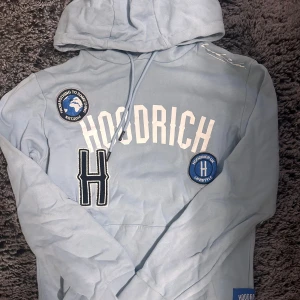 Ljusblå hoodie från Hoodrich - Säljer en ljusblå hoodie från Hoodrich med tryck och broderade detaljer. Den har en stor huva och långa ärmar, perfekt för en avslappnad stil. Hoodien har texten 'HOODRICH' framtill och en stor bokstav 'H'.