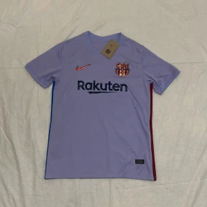 FC Barcelona Fotbollströja - Säljer en lila FC Barcelona fotbollströja från Nike med korta ärmar. Tröjan har klubbens emblem på bröstet och sponsorerna Rakuten och UNICEF tryckta på fram- och baksidan. Snygga detaljer i rött och blått längs sidorna.