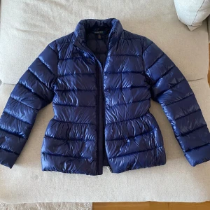Blå dunjacka från Polo Ralph Lauren - Säljer en snygg blå dunjacka från Polo Ralph Lauren. Jackan har en dragkedja framtill och långa ärmar. Perfekt för kyligare dagar med sin pufferdesign som håller värmen. 12-14 år. Nypris ca 3000.