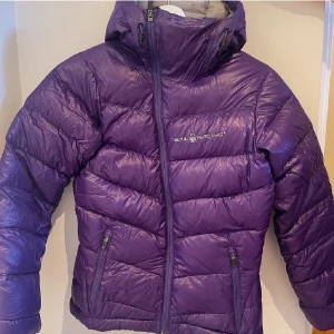 Lila dunjacka från Sail Racing - Snygg lila dunjacka från Sail Racing med dragkedja och hög krage. Perfekt för kyliga dagar med sin pufferdesign och praktiska fickor.