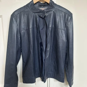 Blå skinnjacka med dragkedja - Snygg blå skinnjacka med texturerad yta och dragkedja framtill. Jackan har en stilren design med långa ärmar och en hög krage med knappdetalj. 