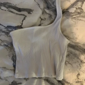 Vit ribbad oneshoulder topp - Snygg vit ribbad topp med oneshoulder-design. Perfekt för en stilren och modern look. Toppen är croppad och passar utmärkt till både jeans och kjol.