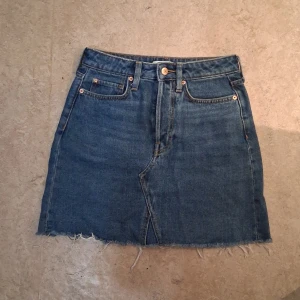 Jeanskjol från H&M - Snygg jeanskjol från H&M i klassisk blå denim. Kjolen har en rå kant nertill och femficksdesign med knapp och dragkedja framtill.