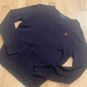 Mörkblå tröja från Polo Ralph Lauren - Säljer en stilren mörkblå tröja från Polo Ralph Lauren i Pima Cotton. Tröjan har en klassisk v-ringning och ett broderat orange märke på bröstet. Perfekt för en avslappnad men elegant look.