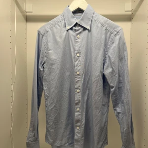 Ljusblå skjorta med knappar - Snygg ljusblå skjorta med klassisk krage och knappar hela vägen ner. Perfekt för en stilren look. Nytvättad och därav det skrynkliga, stryker den på begäran. 