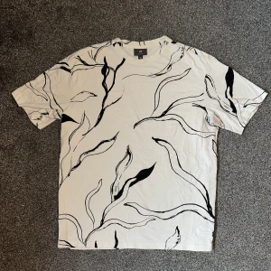 Vit t-shirt med svart mönster från H&M - Säljer en vit t-shirt från H&M med ett abstrakt svart mönster. T-shirten har en normal passform och korta ärmar. Perfekt för en stilren och modern look.