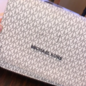 Vit och grå kuvertväska från Michael Kors - Snygg kuvertväska från Michael Kors i vitt och grått med ett stilrent MK-mönster. Väskan har en kedjedetalj och ett rutigt innerfoder. Perfekt för att bära det viktigaste med stil. Använder ej längre. 