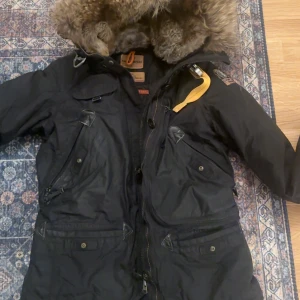 Svart parkajacka med pälsdetaljer - Snygg svart parkajacka från Parajumpers med pälsfodrad huva och flera praktiska fickor. Jackan har dragkedja och knappar framtill samt en quiltad insida för extra värme. Perfekt för kalla dagar.