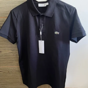 Svart pikétröja från Lacoste - Snygg svart pikétröja från Lacoste med klassisk krage och knappar framtill. Den är oanvänd och har en i storlek M och S. Ny pris 1299kr