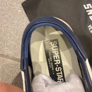 Golden Goose Super-Star sneakers - Snygga vita Golden Goose Super-Star sneakers med grå stjärndetalj och blå häl. Skorna har grå snörning och en klassisk design som passar perfekt för en avslappnad stil. storlek 41. jag har haft dem i ungefär några måndaer fick dem i presnt har ey kvitto tyvärr. priset kan duikteras.hör av er om de nåt 