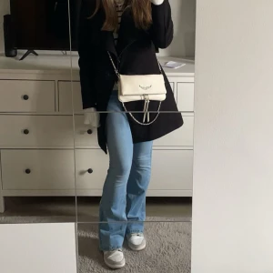 Vit axelväska med kedja - Snygg vit axelväska med kedjedetalj och broderad logga på framsidan. Väskan har en stilren design och passar perfekt för en trendig look. Den är liten och smidig att bära med sig. Köpt i spanien!