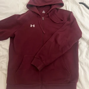 Vinröd hoodie från Under Armour - Säljer en vinröd hoodie från Under Armour med dragkedja och huva. Tröjan har långa ärmar och en liten logotyp på bröstet. Perfekt för en avslappnad stil.