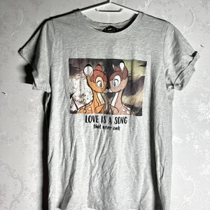 Grå Disney t-shirt med Bambi-tryck - Säljer en grå t-shirt från FB Sister med ett sött Bambi-tryck och texten 'Love is a song that never ends'. Perfekt för Disney-fans! Kortärmad och i mjukt material.