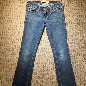 Blå jeans från Hollister - Snygga blå jeans från Hollister med klassisk femficksdesign och guldfärgade sömmar på bakfickorna. De har en lågmidjad passform  och är bootcut. Midja rakt över: 36cm. Lår: 22cm. Innerben: 80cm