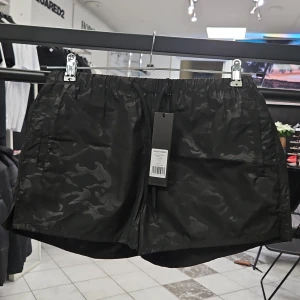 Svarta camouflagemönstrade shorts från Antony Morato - Snygga svarta shorts från Antony Morato med ett diskret camouflagemönster. De har en elastisk midja med snörning för bästa passform. Perfekta för en avslappnad stil. Fickor på sidan och bak. Badshorts storlek S. Material: 94% Polyester, 6% Elastan. Ny