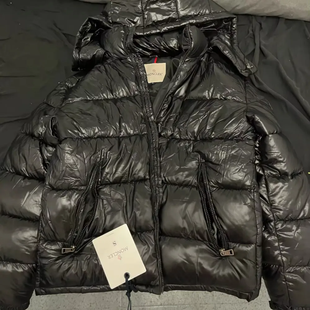 Snygg svart dunjacka från Moncler med huva och dragkedja. Jackan har ett stilrent märke på ärmen och praktiska fickor med dragkedjor. Perfekt för kyliga dagar.. Takit.