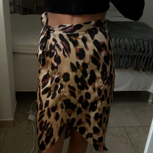 Leopardmönstrad omlottkjol - Trendig leopardmönstrad omlottkjol i beige och svart med knytning i midjan. Kjolen har en smickrande passform och är perfekt för att ge en djärv touch till din outfit.