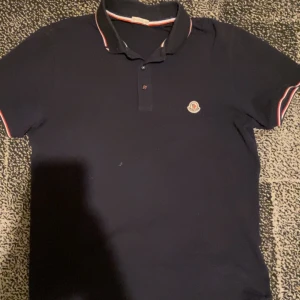 Moncler piké - Säljer en snygg mörkblå pikétröja från Moncler med klassisk krage och knappar. Tröjan har en broderad logotyp på bröstet och detaljer i rött och vitt på kragen och ärmsluten. NFC fungerar