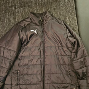 Svart pufferjacka från Puma - Säljer en svart pufferjacka från Puma med dragkedja och broderad logga på bröstet. Jackan har långa ärmar och är perfekt för kyligare väder. 