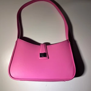 Rosa axelväska i syntet - Snygg rosa axelväska i syntetmaterial med en stilren design. Väskan har en praktisk dragkedja och ett elegant spänne framtill. Perfekt för att ge en färgklick till din outfit!