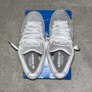 Grå Adidas sneakers - Snygga grå Adidas sneakers i mocka med vita detaljer och snörning. De har en klassisk design med tre ränder på sidan och en gummisula i brunt. Perfekta för en avslappnad stil. De är sprayade med skyddspray vilket hjälper mot snö, regn och smuts. Helt äkta!!!