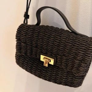 Svart axelväska med gulddetalj - Snygg svart axelväska i flätat material med en elegant guldfärgad låsdetalj. Perfekt för att bära dina nödvändigheter med stil. Väskan har en justerbar axelrem för bekväm passform.