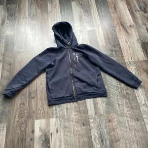 Mörkblå hoodie med dragkedja - Säljer en stilren mörkblå hoodie med dragkedja och justerbar huva. Perfekt för en avslappnad look. Den har långa ärmar och en praktisk ficka framtill. Passar bra till både jeans och träningsbyxor.