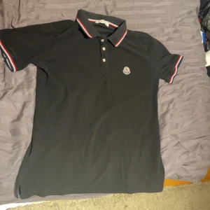 Svart pikétröja från Moncler - Snygg svart pikétröja från Moncler med klassisk krage och knappar. Tröjan har röda och vita detaljer på kragen och ärmsluten samt Monclers logotyp broderad på bröstet. Perfekt för en stilren look. Säljs pga att den är för liten