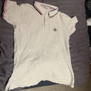 Vit pikétröja från Moncler - Säljer en klassisk vit pikétröja från Moncler med broderad logga på bröstet. Tröjan har en krage med röd och blå rand samt korta ärmar med matchande ränder. Perfekt för en stilren look. Säljs pga att den har en liten fläck på bröstet samt att den är för liten 