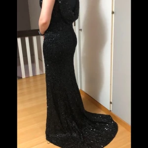 Svart glittrig långklänning - Elegant svart långklänning med glittrande detaljer. Klänningen har en figurnära passform och en vacker drapering i ryggen. Perfekt för en glamorös kväll.