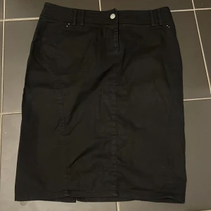 Svart kjol med dödskallemotiv - Snygg svart kjol med ett coolt dödskallemotiv i strass på baksidan. Kjolen har en klassisk design med knäppning fram och bälteshällor. Perfekt för att ge din outfit en edgy touch.