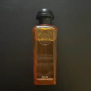 Eau de mandarine ambrée parfym - Elegant parfymflaska med en varm orange ton och svart lock. Doften är fräsch och citrusaktig med inslag av mandarin och bärnsten. Perfekt för den som älskar en lätt och uppfriskande doft. 100 ml 