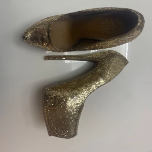 Glittriga guldiga pumps - Snygga pumps i glittrigt guld med hög klack och spetsig tå. Perfekta för att lysa upp vilken outfit som helst. Dessa skor är ett måste för den som vill sticka ut.