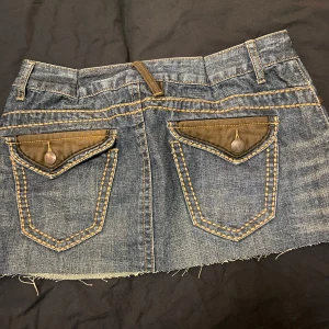 Jeanskjol från HoneyBblue - Snygg lågmidjad jeanskjol från HoneyBblue i 100% bomull. Kjolen har en kort längd med rå kant och dekorativa bakfickor med knappar. Den har en klassisk jeanslook med kontrastsömmar och bälteshällor.