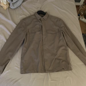 Beige overshirt från H&M - Säljer en stilren beige overshirt från H&M. Skjortan har långa ärmar och två praktiska bröstfickor med lock. Perfekt för lager-på-lager-stil och passar till många olika outfits.