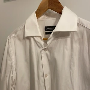 Vit skjorta från Dressmann - Säljer en stilren vit skjorta från Dressmann i slim fit. Skjortan är tillverkad i bomull med stretch och har långa ärmar samt knappar framtill. Perfekt för en elegant look. Den är använd 1 gång på bröllop! Köpt för 600kr! 