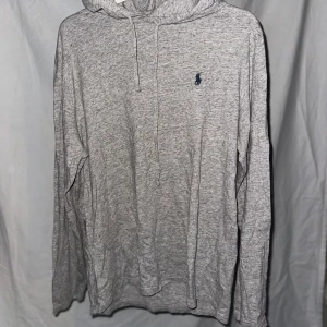 Grå hoodie från Ralph Lauren - Säljer en stilren grå hoodie från Ralph Lauren med det klassiska logotypen broderad på bröstet. Tröjan har långa ärmar och en justerbar huva med dragsko. Perfekt för en avslappnad look.