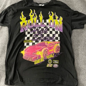 Svart t-shirt med biltryck från FB Sister - Cool svart t-shirt från FB Sister med ett färgglatt tryck av en sportbil och texten 'Roll On High Speed'. T-shirten har korta ärmar och ett rutigt mönster i bakgrunden av trycket.