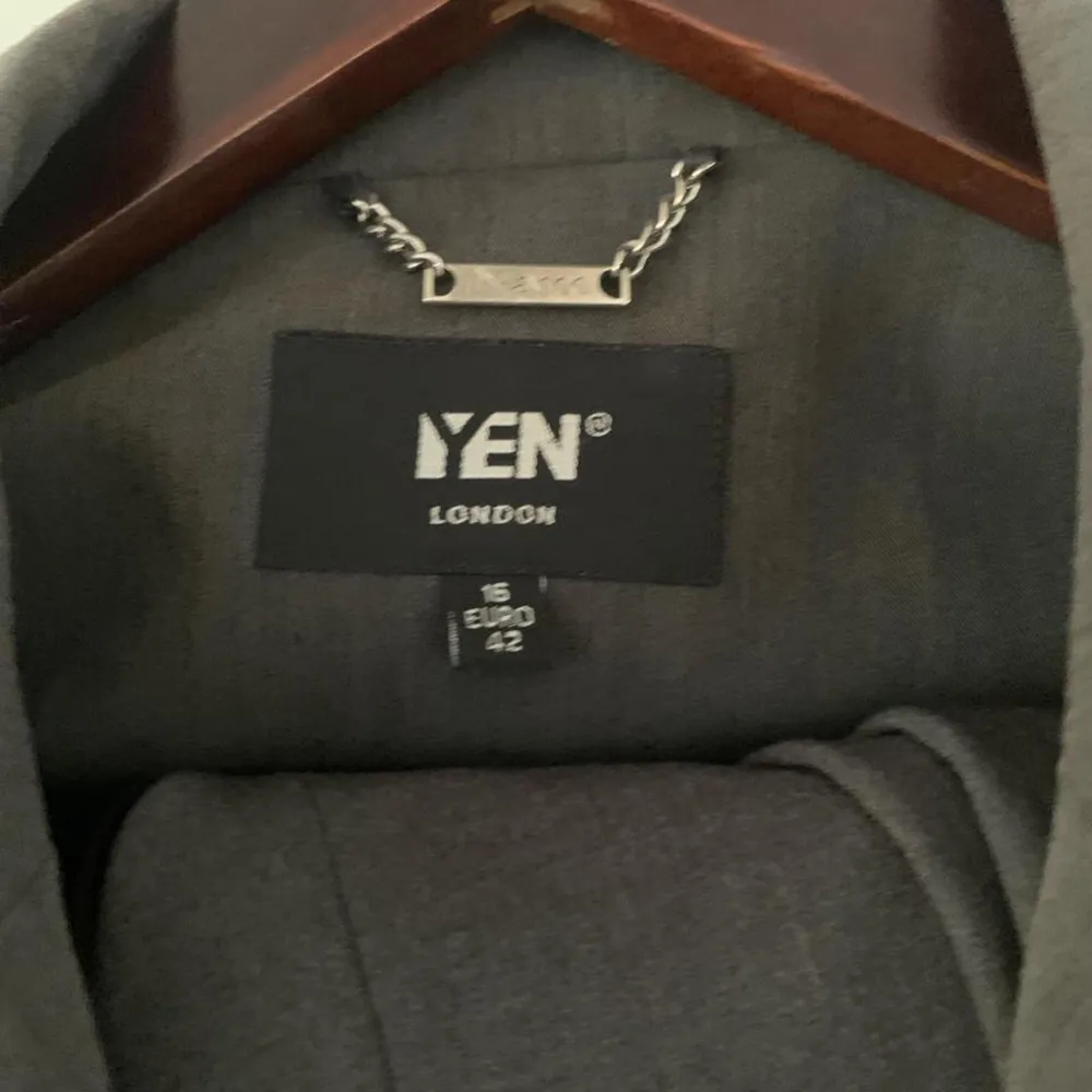 Elegant grå kavaj från YEN London med knytband i midjan och en knapp framtill. Perfekt för en stilren look. Kavajen har en klassisk krage och långa ärmar. Storlek 42. Puvut & Bleiserit.