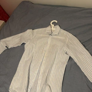 Vit och blå randig skjorta från Massimo Dutti - Säljer en stilren vit skjorta med blå ränder från Massimo Dutti. Skjortan är tillverkad i en skön blandning av bomull och linne, perfekt för en avslappnad men ändå uppklädd look. Den har långa ärmar och knappar framtill. Denna pärlan från Massimo dutti, kan kombineras med den bruna tröjan jag säljer som funkar perfekt som en fräsch sommar look! Nu börjar solen smyga in så det passar perfekt! Använt 1 gång, nypris 1000