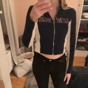 Marinblå och vit croppad tröja med dragkedja - Säljer en marinblå och vit croppad tröja med dragkedja och texten 'New York' på bröstet. Tröjan har långa ärmar, luva och en sportig look. Använd 5-10 gånger 