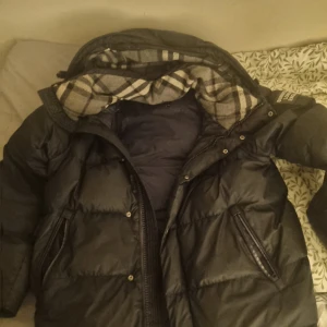 Svart dunjacka från Burberry - Säljer en snygg svart dunjacka från Burberry med klassiskt rutigt foder. Jackan har dragkedja och knappar framtill samt en hög krage för extra värme. Perfekt för kyliga dagar!