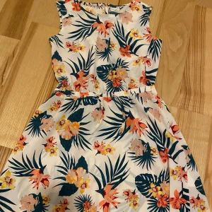 Blommig klänning  - Säljer en somrig blommig klänning från Basic Needs. Klänningen är ärmlös och har ett färgglatt mönster med blommor i orange, rosa och grönt på en vit bakgrund. Perfekt examensklänning! 🌸 