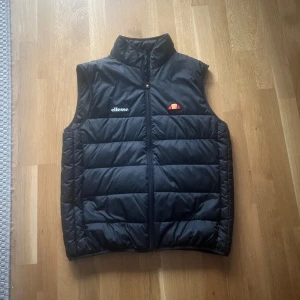 Svart dunväst från Ellesse - Snygg svart dunväst från Ellesse med märkets logga på bröstet. Västen har en hög krage och är perfekt för kyligare dagar. Den quiltade designen ger både stil och värme.