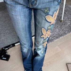 Vintage jeans med broderi - Snygga vintage jeans med unikt blommigt broderi längs benet och en blomma på bakfickan. De har en lågmidjad passform. Perfekta för att ge din outfit en färgglad touch! Det ska vara färg på blommorna men dessa är köpta i en secondhand butik och då var de redan så. Knappen på jeansen är även utbytt till en mycket snyggare knapp. 💞Midjemått: 32cm (mätt platt) Höftbredd: ca 40cm (mätt platt) Grenmått: 21cm  Lårbensmått: 22cm (mätt platt) Innerbenslängd: 81cm