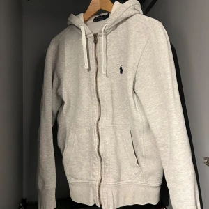 Grå hoodie från Ralph Lauren - Säljer en stilren grå hoodie från Ralph Lauren med dragkedja och huva. Tröjan har långa ärmar och en liten broderad logga på bröstet. Perfekt för en avslappnad stil. Skriv vid fler frågor och funderingar👍🏽