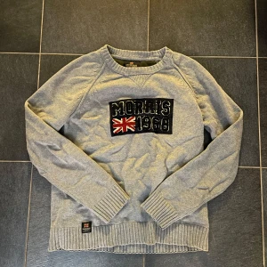 Grå tröja från Morris - Säljer en stilren grå tröja från Morris med ett broderat märke på bröstet som inkluderar en brittisk flagga och texten 'Morris 1968'. Tröjan har långa ärmar och ribbade muddar vid ärmslut och nederkant. Skriv vid funderingar 