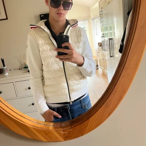 Vit quiltad jacka från Moncler - Säljer en snygg vit quiltad jacka från Moncler med huva och dragkedja. Jackan har långa ärmar och en stilren design med röd och svart randdetalj på kragen. Perfekt för kyligare dagar!